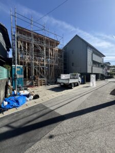 Construction site on a residential street with timber framing and metal scaffolding beside a grey building; materials and a small truck nearby.