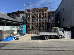 Construction site with a wooden-framed building surrounded by metal scaffolding between two existing buildings; a blue portable toilet and construction tools visible in the foreground.
