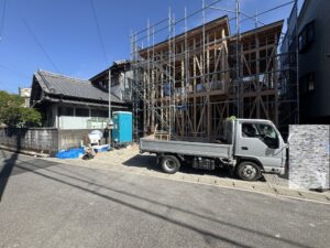Wood-framed building under construction surrounded by metal scaffolding beside a traditional tiled house on a sunny street